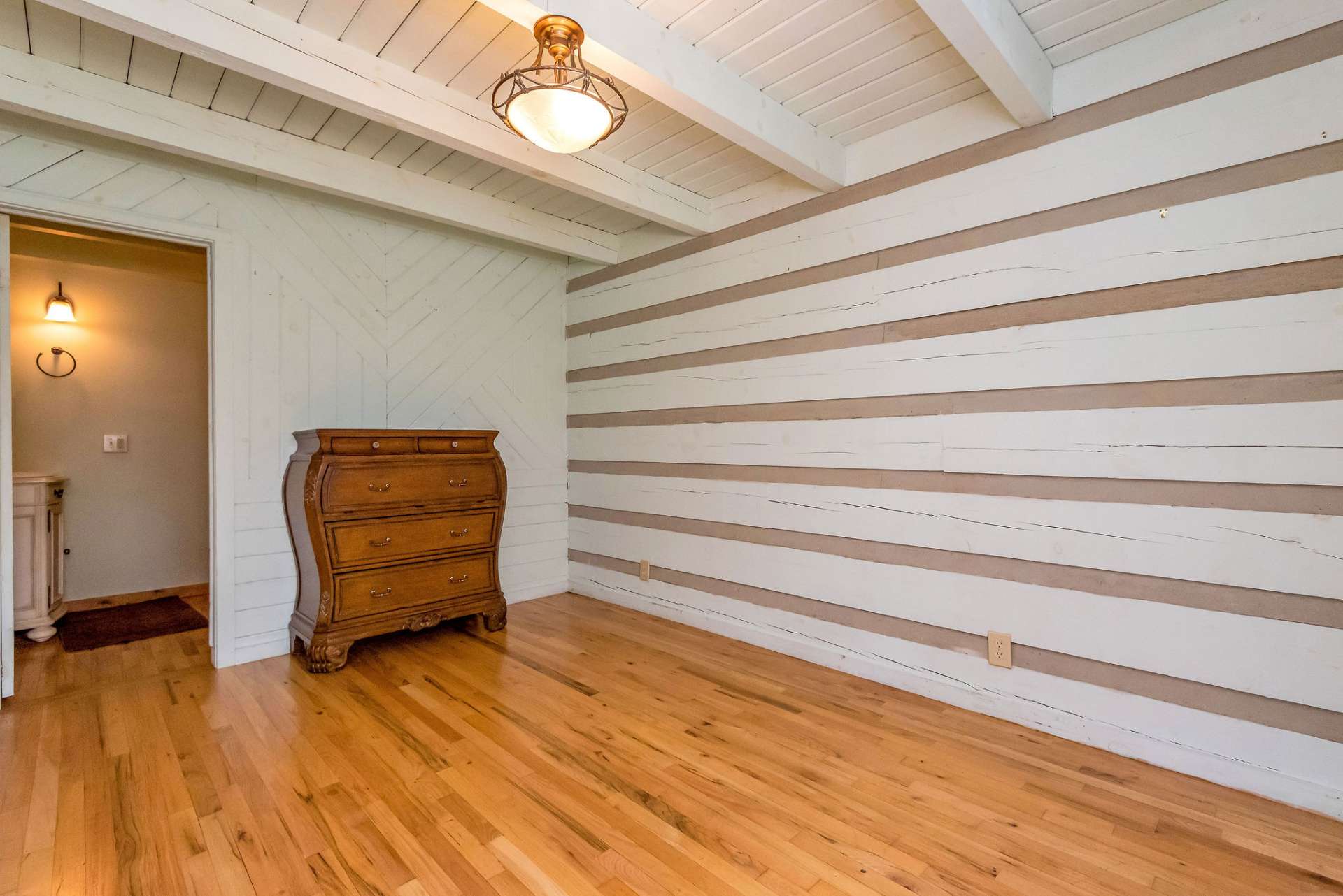 The primary bedroom, located on the main level, features log walls, ceiling, and exposed beams that have been painted to brighten the room, adding a touch of modernity while preserving its rustic appeal.